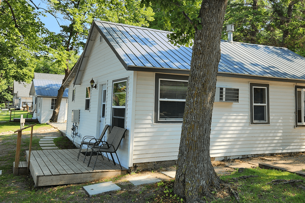 White siding on one story cabin with an ongoing row of cabins with trees and lake in distance